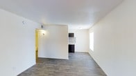 This single-floor unit features an open, functional layout designed to maximize the available living space. The main living area is finished with wood-look plank flooring in a cool gray tone, lending a modern, clean aesthetic throughout. Bright white walls and a smooth white ceiling enhance the sense of openness and allow natural light, entering through a window on the far wall, to reflect evenly across the space.

A partial dividing wall separates the living area from the rest of the unit, creating a subtle visual boundary without fully closing off the floor plan. Beyond this partition, a compact kitchen comes into view, featuring dark cabinetry paired with a mosaic tile backsplash that adds a decorative accent. A microwave is mounted above the counter, making efficient use of vertical space.

A hallway visible to the left, lit by a wall-mounted light fixture, leads to additional areas of the unit. The overall palette of the space—neutral walls, gray flooring, and dark kitchen finishes—creates a cohesive, contemporary look. The single-floor layout ensures easy accessibility and seamless flow between the living and kitchen areas.