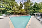 This outdoor amenity space centers around a generously sized swimming pool with clear turquoise water, offering a resort-like atmosphere ideal for relaxation and recreation. The pool features a gradually varying depth, clearly marked along the edges, and is accessible via built-in entry steps with handrails. Surrounding the pool is a broad, well-maintained concrete sun deck furnished with an extensive row of lounge chairs and chaise lounges positioned along multiple sides, providing ample space for sunbathing. Several market umbrellas are strategically placed throughout the deck, offering shaded seating options for those seeking refuge from the sun. A decorative pergola structure anchors one corner of the deck, adding an architectural focal point and additional shaded gathering space. The entire pool area is enclosed by a security fence and is enveloped by a dense backdrop of mature trees, including tall pines and flowering crepe myrtles, creating a private, natural setting. Adjacent to the pool is a well-appointed single-story clubhouse building with a classic pitched roof, dormer windows, white siding, and large windows that face the pool deck. The building's covered porch connects directly to the pool area, providing a functional transition space. Together, these elements create a cohesive, inviting outdoor environment designed for community enjoyment.