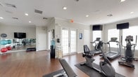 This single-floor fitness center offers a comprehensive workout environment designed to accommodate a variety of exercise routines. The space is divided into distinct functional zones, each thoughtfully outfitted with equipment and amenities. The cardio area houses multiple treadmills, an elliptical, and a recumbent bike, all positioned on protective rubber mats atop rich hardwood flooring. A wall-mounted flat-screen TV provides entertainment during workouts.

The strength training section features a multi-station cable machine alongside a full dumbbell rack ranging across multiple weight increments, complemented by a large framed mirror that enhances the sense of space and allows for form monitoring. Additional free weights and a flat workout bench are also available.

A separate open-floor area serves as a dedicated aerobic and stretching zone, equipped with stability balls, a step platform, and a wall of floor-to-ceiling mirrors. Crown molding and neutral gray-white walls lend a polished, clean aesthetic throughout. French doors with frosted glass panels provide natural light while maintaining privacy, and windows along multiple walls further brighten the interior. A water cooler station and recessed lighting complete the practical, well-appointed environment.