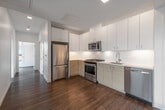 This modern single-floor residence showcases a thoughtfully designed open layout with warm hardwood flooring throughout. The kitchen is a standout feature, equipped with stainless steel appliances including a gas range, built-in microwave, refrigerator, and dishwasher. Flat-front cabinetry in a light wood finish extends to the ceiling, complemented by white upper cabinets and light quartz countertops, creating a clean, contemporary aesthetic. A hallway connects the kitchen to the remaining rooms, maintaining an efficient flow throughout the space.

The living area is defined by striking floor-to-ceiling corner windows that frame panoramic urban views, including a notable historic building in the cityscape, flooding the space with natural light. The bedroom features its own large window with horizontal blinds and direct access to a walk-in closet, offering both comfort and storage.

The bathroom is finished with distinctive terracotta hexagonal floor tiles, a wood-toned vanity with integrated sink, a full soaking tub with tile surround, and a wall-mounted medicine cabinet. A washer and dryer unit is also included within the layout. White walls throughout provide a neutral, contemporary backdrop that enhances the bright, airy character of the entire space.