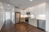 This modern single-floor residence showcases a thoughtfully designed open layout with warm hardwood flooring throughout. The kitchen is a standout feature, equipped with stainless steel appliances including a gas range, built-in microwave, refrigerator, and dishwasher. Flat-front cabinetry in a light wood finish extends to the ceiling, complemented by white upper cabinets and light quartz countertops, creating a clean, contemporary aesthetic. A hallway connects the kitchen to the remaining rooms, maintaining an efficient flow throughout the space.

The living area is defined by striking floor-to-ceiling corner windows that frame panoramic urban views, including a notable historic building in the cityscape, flooding the space with natural light. The bedroom features its own large window with horizontal blinds and direct access to a walk-in closet, offering both comfort and storage.

The bathroom is finished with distinctive terracotta hexagonal floor tiles, a wood-toned vanity with integrated sink, a full soaking tub with tile surround, and a wall-mounted medicine cabinet. A washer and dryer unit is also included within the layout. White walls throughout provide a neutral, contemporary backdrop that enhances the bright, airy character of the entire space.