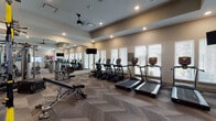 This fitness center offers a comprehensive workout environment designed to accommodate a wide range of exercise needs. The space is laid out across a single floor and features a distinctive herringbone-patterned carpet throughout the main workout area, lending a modern aesthetic to the room. Recessed lighting and ceiling fans ensure the space remains well-lit and comfortable during use.

The cardio zone is well-appointed with multiple True-brand treadmills and elliptical machines arranged in rows along a windowed wall, where slatted blinds allow natural light to filter in while maintaining privacy. Wall-mounted flat-screen televisions are positioned for easy viewing from the cardio equipment.

A dedicated strength and functional training area features a multi-rack system stocked with medicine balls, resistance bands, TRX suspension trainers, and a full set of dumbbells. An adjustable weight bench and cable machine expand the range of available exercises. A large mirror spans one wall, enhancing the sense of space and allowing users to monitor their form.

A separate area with hardwood-style flooring houses spin bikes and additional equipment, providing a distinct zone for cycling workouts. Colorful artwork accents the walls, adding a vibrant touch to the clean, neutral color palette of warm whites and soft grays that defines the overall ambiance of this well-rounded fitness facility.