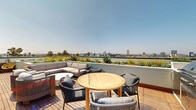 Located in the heart of Los Angeles, this rooftop terrace at 360 N Hayworth Ave offers an expansive open-air retreat with sweeping panoramic views of the Los Angeles skyline and surrounding neighborhoods, including glimpses of iconic palm tree-lined streets and distant high-rises. The deck is finished with warm hardwood planking throughout, lending a refined, resort-like aesthetic to the entire space.

The terrace is thoughtfully organized into multiple distinct zones for lounging, dining, and entertaining. A generously sized sectional sofa anchors one of the primary lounge areas, complemented by round accent chairs with woven rope detailing and a circular white coffee table. Additional seating vignettes — including armchairs and bistro-height tables — are distributed across the deck, accommodating both intimate gatherings and larger social events.

A dedicated outdoor kitchen and grilling station provide functional amenities for al fresco entertaining, positioned alongside a dining table and chairs ideal for open-air meals. A striking dark navy, vertically ribbed metal architectural wall serves as both a dramatic design element and a functional divider, adding a bold contemporary contrast to the natural wood and neutral furnishings.

Lush planted borders with succulents and ornamental grasses frame the perimeter, integrating greenery into the urban setting. Under the abundant Southern California sunshine, this rooftop terrace delivers a versatile and sophisticated outdoor experience.