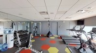 The fitness center at 120 N Pearl St in Port Chester, NY is a thoughtfully equipped facility designed to support a comprehensive workout experience. The space features a distinctive flooring design with bold red and yellow hexagonal patterns set against a neutral gray carpet, adding a dynamic, energetic aesthetic to the room.

Floor-to-ceiling mirrors line the walls, visually expanding the space and providing users with proper form feedback during exercises. The suspended tile ceiling houses recessed lighting that ensures the area is bright and well-illuminated throughout.

The cardio section is well-stocked with True treadmills, a Concept2 rowing machine, spin bikes, elliptical trainers, and a recumbent bike, with wall-mounted flat-screen televisions positioned for easy viewing during workouts. The strength training area includes a dumbbell rack with a full range of free weights, a weight bench, and a cable pulley machine. Functional training accessories such as medicine balls on a dedicated rack, battle rope equipment, and a speed bag round out the offerings.

Natural light filters into the space through large windows, and a staircase with glass railings connects to adjacent areas of the building. The overall ambiance is modern, clean, and motivating, making this fitness center a capable and inviting destination for residents and users alike.