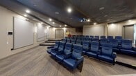 This dedicated screening room delivers an authentic cinema experience within a single-floor layout. The main theater space is anchored by multiple rows of plush, blue leather seats arranged in a tiered, stadium-style configuration, each equipped with integrated cup holders for added convenience. Seating faces a large projection screen mounted at the front of the room, paired with a ceiling-mounted projector that ensures a sharp, immersive viewing experience. Recessed lighting and a drop ceiling with a dark finish create a moody, theater-appropriate ambiance throughout.

A concession area along one wall features dark wood cabinetry, a countertop with a sink, and a beverage cooler beneath — all anchored by a standout classic red popcorn cart on yellow wheels, adding a whimsical, nostalgic touch. Framed movie-themed artwork adorns the neutral-toned walls, reinforcing the cinematic atmosphere. Patterned carpeting in warm gray tones runs throughout the space, while accessibility is thoughtfully addressed with handrails along a side ramp. Additional doors provide convenient entry and exit points. The overall design balances functional theater infrastructure with welcoming, polished finishes, making it well-suited for both entertainment and group viewing events.