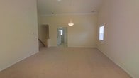 This single-floor home offers an open and spacious layout designed to maximize comfort and functionality. The main living area is immediately notable for its soaring ceilings, which lend a grand sense of volume and openness to the space. Soft, neutral-toned wall-to-wall carpeting runs throughout, providing a warm and comfortable underfoot feel while complementing the light, cream-colored walls.

A decorative pendant light fixture serves as a refined focal point, hanging gracefully from the elevated ceiling and adding a touch of elegance to the open living and dining area. Natural light filters in through windows fitted with blinds, brightening the interior and creating an airy atmosphere throughout the day.

Crown molding details along the upper walls add a subtle architectural refinement to the space. A half-wall partition with a stepped countertop defines a transition between areas, providing visual separation while maintaining the open flow of the floor plan. A doorway leads into an adjacent room, hinting at further connected living spaces beyond, while additional hallway access suggests a well-connected and thoughtfully arranged interior. Overall, the space presents a clean, neutral canvas well-suited for a variety of living arrangements.