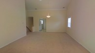 This single-floor home offers an open and spacious layout designed to maximize comfort and functionality. The main living area is immediately notable for its soaring ceilings, which lend a grand sense of volume and openness to the space. Soft, neutral-toned wall-to-wall carpeting runs throughout, providing a warm and comfortable underfoot feel while complementing the light, cream-colored walls.

A decorative pendant light fixture serves as a refined focal point, hanging gracefully from the elevated ceiling and adding a touch of elegance to the open living and dining area. Natural light filters in through windows fitted with blinds, brightening the interior and creating an airy atmosphere throughout the day.

Crown molding details along the upper walls add a subtle architectural refinement to the space. A half-wall partition with a stepped countertop defines a transition between areas, providing visual separation while maintaining the open flow of the floor plan. A doorway leads into an adjacent room, hinting at further connected living spaces beyond, while additional hallway access suggests a well-connected and thoughtfully arranged interior. Overall, the space presents a clean, neutral canvas well-suited for a variety of living arrangements.