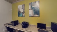 This single-floor business center is designed to meet essential productivity needs in a compact, well-organized setting. The space features a continuous countertop workstation that runs along the wall, providing ample surface area for computer workstations and office equipment, including a multifunction printer/scanner and desktop computer monitors with keyboards readily accessible for guest or tenant use.

The walls are painted in a warm yellow-green tone, adding a distinctive and energizing character to the space. Two large abstract canvas paintings hang prominently above the workstation, incorporating a palette of teal, grey, and white that complements the wall color and adds a polished, contemporary aesthetic to the functional environment.

The layout is straightforward and efficient, prioritizing ease of use and accessibility. The countertop setup ensures that multiple users can work simultaneously without crowding. Overall, the business center strikes a balance between practicality and thoughtful design, offering a welcoming atmosphere suited for tasks such as printing, scanning, browsing, and general computer use.
