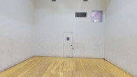 This space is a single, self-contained racquetball court designed to meet the functional demands of both recreational and competitive play. The court is enclosed on all sides by tall, smooth white walls that serve as active playing surfaces, showing the characteristic wear marks typical of a well-used court. The floor is constructed of traditional hardwood planking finished in a natural tone, with clearly marked red boundary lines including service boxes, service lines, and a dashed short line — all consistent with standard racquetball court specifications. Overhead, flush-mounted skylight-style lighting panels provide even, diffused illumination across the entire playing surface. The rear wall features a solid door with a small viewing window, alongside a ventilation grille and an additional observation window positioned near the top of the wall, allowing spectators or coaches to monitor play from outside. The tall ceiling height accommodates the full range of shots required by the sport, including ceiling shots. The overall environment is utilitarian and purpose-built, prioritizing performance and functionality. The enclosed design ensures excellent ball rebound consistency, making this court well-suited for singles or doubles racquetball.