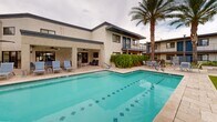 Located in Phoenix, AZ, this outdoor pool area offers a resort-inspired retreat designed for relaxation and recreation. The centerpiece is a generously sized rectangular swimming pool with vivid turquoise water and decorative tiled accents along the floor, creating a visually striking focal point. The surrounding deck is finished in neutral stone pavers, providing a clean and cohesive aesthetic throughout the space.

Ample seating is available poolside, including rows of sleek lounge chairs paired with striped market umbrellas that offer shade on sunny Arizona days. Multiple seating zones are distributed around the pool, including areas beneath a structured pergola and a covered patio equipped with ceiling fans — ideal for enjoying the outdoors while staying cool.

The landscaping is thoughtfully curated, featuring tall, mature palm trees that frame the space and add a tropical character, alongside ornamental shrubs, flowering plants, and a decorative accent tree that provides visual softness against the stucco building exteriors. The surrounding two-story buildings with balconies create an intimate, courtyard-like atmosphere that encloses the pool area while maintaining an open-air feel. This outdoor space is well-suited for both leisurely lounging and social gatherings in the heart of Phoenix.