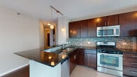 This thoughtfully designed high-rise residence offers a seamless blend of modern finishes and urban living. The open-concept kitchen is a standout feature, showcasing rich dark wood cabinetry extending to the ceiling, dramatic dark granite countertops, a mosaic tile backsplash in muted blues and greens, and a full suite of stainless steel appliances including a gas range, built-in microwave, and refrigerator. A peninsula with pendant lighting above provides additional prep space and casual seating. The spacious living area is defined by gleaming hardwood floors and an expansive wall of floor-to-ceiling windows that frame sweeping city skyline views, filling the room with abundant natural light. The bedrooms are carpeted for comfort and feature generous walk-in closets with built-in shelving. Corner bedroom windows offer dramatic panoramic views of the urban skyline. The bathrooms are finished with large-format tile, dark-toned vanities topped with granite, a soaking tub, and a separate glass-enclosed walk-in shower. Track lighting and thoughtful fixtures throughout complete the clean, contemporary aesthetic of this well-appointed urban residence.