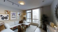This single-floor urban residence offers a thoughtfully designed living space that balances comfort and contemporary style. The living area serves as the heart of the home, anchored by large floor-to-ceiling windows dressed with sheer white curtains that invite an abundance of natural light while framing views of the surrounding cityscape. The room features warm hardwood flooring that complements the neutral, earthy color palette throughout. A plush white sectional sofa is paired with rustic wood stump coffee tables, creating a casual yet curated atmosphere. Track lighting and a flush-mount ceiling fixture provide layered illumination, enhancing the room's versatility. A tall floor lamp adds a soft, ambient glow alongside the natural light. Decorative framed artwork adorns the walls, lending a personalized, gallery-like touch. A lush indoor plant in the corner brings a natural, organic element to the interior. The clean, modern lines of the architecture are complemented by warm décor accents, creating an inviting and functional living environment that feels both polished and livable — well-suited for both relaxed daily living and entertaining.