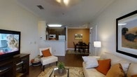 This single-floor space features an open-concept design that seamlessly connects the living room, dining area, and kitchen. The living room is anchored by a neutral-toned sofa and accent chair, both styled with orange throw pillows that complement the warm color palette throughout. A glass-topped coffee table and a patterned area rug add texture and definition to the seating area. Hardwood-style flooring extends throughout, lending a cohesive and polished look to the space.

The kitchen is positioned behind a half-wall partition with decorative columns, offering a semi-open feel while maintaining a degree of separation. It features dark cabinetry, a white refrigerator, microwave, and under-cabinet lighting that brightens the workspace. A pass-through ledge connects the kitchen to the living area, ideal for casual interaction.

The dining area is set apart by a rich, warm orange accent wall that serves as a striking focal point. A dark wood dining table surrounded by matching chairs sits atop an area rug, completing the space. Crown molding and neutral wall tones throughout add a refined finishing touch to the overall ambiance.