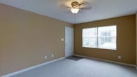 This single-floor residential space offers a clean and functional layout designed for comfortable everyday living. The main room is finished with wall-to-wall carpeting in a neutral tone, creating a warm and inviting atmosphere underfoot. The walls are painted in a soft, earthy golden-tan hue that adds warmth and depth to the interior without overwhelming the senses.

A ceiling fan with a decorative brass-toned fixture and integrated light sits centrally overhead, providing both air circulation and ambient lighting to the space. White baseboards run along the perimeter of the room, offering a crisp contrast to the warm wall color and contributing to a tidy, well-maintained aesthetic.

Natural light enters generously through a large window, which looks out toward an exterior view, brightening the room throughout the day. A white entry door with a small welcome mat positioned at its base provides a clear and welcoming point of entry. Electrical outlets are thoughtfully placed along the walls, adding to the practicality of the room.

Overall, the space presents a straightforward, move-in-ready environment with a modest but comfortable character, suitable for a variety of living arrangements.
