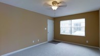 This single-floor residential space offers a clean and functional layout designed for comfortable everyday living. The main room is finished with wall-to-wall carpeting in a neutral tone, creating a warm and inviting atmosphere underfoot. The walls are painted in a soft, earthy golden-tan hue that adds warmth and depth to the interior without overwhelming the senses.

A ceiling fan with a decorative brass-toned fixture and integrated light sits centrally overhead, providing both air circulation and ambient lighting to the space. White baseboards run along the perimeter of the room, offering a crisp contrast to the warm wall color and contributing to a tidy, well-maintained aesthetic.

Natural light enters generously through a large window, which looks out toward an exterior view, brightening the room throughout the day. A white entry door with a small welcome mat positioned at its base provides a clear and welcoming point of entry. Electrical outlets are thoughtfully placed along the walls, adding to the practicality of the room.

Overall, the space presents a straightforward, move-in-ready environment with a modest but comfortable character, suitable for a variety of living arrangements.