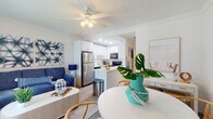 Located on Spanish River Road in Boca Raton, Florida, this single-floor residence embraces a relaxed coastal aesthetic throughout its thoughtfully designed layout. The open-plan living, dining, and kitchen area forms the heart of the home, unified by light wood-tone flooring and crisp white walls that enhance the sense of brightness and airiness. A ceiling fan overhead adds both function and a casual, breezy feel to the living space, which features a large window offering views of lush palm trees outside.

The kitchen is efficiently designed with white cabinetry, quartz-style countertops, a stainless steel refrigerator, and black appliances including an electric range with an over-the-range microwave. A peninsula provides a natural division between the kitchen and the living area. The dining space accommodates a round white table with rattan-style chairs, complemented by a wall-mounted television and a white sideboard with woven basket storage.

The bedroom is comfortably scaled, featuring a ceiling fan, a large built-in wardrobe with ample storage, and a window that brings in natural light and outdoor views. The bathroom presents a clean, modern finish with white cabinetry, quartz countertop, an undermount sink with brushed nickel fixtures, and a round mirror. Throughout, the coastal-inspired décor creates a cohesive, welcoming ambiance.