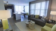 This single-floor residence showcases a thoughtfully designed open-concept layout that seamlessly integrates the living, dining, and kitchen areas into one cohesive, flowing space. Large floor-to-ceiling windows with sleek horizontal blinds line the exterior walls, flooding the interior with abundant natural light and offering views of the surrounding environment.

The living area is anchored by a contemporary charcoal sofa accented with plaid and solid green throw pillows, paired with a distinctive natural wood stump coffee table that adds an organic, earthy contrast to the otherwise modern aesthetic. A neutral woven area rug defines the seating zone underfoot.

Adjacent to the living area, the dining space features a clean-lined table surrounded by upholstered green chairs, positioned directly beneath a pendant light fixture. The kitchen extends naturally from the dining area, highlighted by a white island with a built-in sink, dark lower cabinetry, and additional pendant lighting overhead.

Dark hardwood flooring runs consistently throughout the space, grounding the neutral and green color palette. The overall ambiance is contemporary and inviting, balancing modern finishes with warm, natural accents.