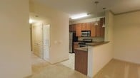This single-floor unit is thoughtfully designed to maximize functionality and flow. The kitchen serves as a central feature, showcasing rich medium-toned wood cabinetry that extends to the upper walls, providing generous storage throughout. Stainless steel appliances, including a refrigerator, electric range, and over-the-range microwave, lend a modern, practical appeal to the space. A peninsula with a dark countertop and breakfast bar overhang extends outward, creating a natural divide between the kitchen and the adjoining living area while offering additional seating and prep space. Pendant lights hang above the peninsula, adding focused illumination and a decorative touch. The kitchen is further lit by a flush fluorescent ceiling fixture, ensuring a bright, well-lit workspace. The adjacent hallway connects to what appears to be closet or storage space, with double doors providing easy access. Neutral-toned carpet and light-colored walls throughout contribute to a clean, understated aesthetic, while the open layout creates a cohesive and welcoming atmosphere.
