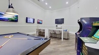 This single-floor amenity lounge is thoughtfully designed to serve as a social and recreational hub. The space is divided into distinct functional zones, each contributing to an inviting and modern atmosphere. Light-toned hardwood-style flooring runs throughout, complementing the crisp white walls accented by classic chair-rail molding and substantial baseboards.

The game area is anchored by a full-size pool table positioned beneath an industrial-style pendant light fixture, with soaring ceilings adding a dramatic sense of height. A large window floods this zone with natural light and offers views to an outdoor courtyard. Locally inspired wall art referencing Deep Ellum, Texas, adds a distinctive sense of place.

Adjacent to the game area is a lounge zone furnished with generous sectional seating, patterned area rugs, and multiple wall-mounted flat-screen televisions paired with soundbars, creating an ideal setting for group viewing or casual socializing. High-top tables with upholstered bar stools provide additional seating alongside dedicated TV screens.

The kitchen area features white shaker cabinetry, striking dark marble-look countertops with a stainless steel undermount sink, stainless steel appliances including a French door refrigerator and built-in microwave, and recessed lighting. A small dining table near the window completes this functional space.
