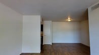 This single-floor space offers a clean and functional layout designed for comfortable everyday living. The main living area is characterized by smooth white walls and dark hardwood-style flooring that runs throughout, creating a warm yet contemporary aesthetic. The open layout allows for flexible furniture arrangements and provides a spacious feel. A partial wall divides the living area from what appears to be a kitchen space, where glimpses of cabinetry and countertop surfaces are visible, suggesting a practical and efficiently designed cooking area. A white interior door leads to an additional room, contributing to the overall sense of connectivity within the unit. Ceiling-mounted lighting provides consistent illumination throughout the space, complementing the bright, neutral color palette of the walls. The minimal, uncluttered design gives the space a fresh and move-in-ready quality. Overall, the space presents a straightforward and functional interior with durable finishes and a cohesive design scheme well-suited for a variety of uses.