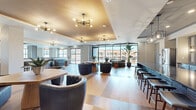 This generously proportioned single-floor amenity space offers a sophisticated and welcoming environment designed for both social gatherings and relaxed lounging. The open-concept layout is anchored by wide-plank hardwood flooring that flows seamlessly throughout, unifying the various functional zones within the space.

The main lounge area is defined by layered tray ceilings with warm gold accents and a series of modern sputnik-style chandeliers that cast an inviting glow. Floor-to-ceiling curtain-wall windows and sliding glass doors bathe the interior in abundant natural light while providing direct access to an outdoor terrace overlooking the surrounding community. Multiple seating arrangements — including tufted leather chairs, curved sectionals, and banquette-style sofas — create distinct conversation areas suited for various group sizes.

A fully equipped catering kitchen features stainless steel appliances, a large stone-topped island, decorative tile backsplash panels, and a wine refrigerator, making the space ideal for hosted events. A bar counter with bar-height seating adds a social focal point nearby.

Additional areas include a corridor with elegant wainscoting and curated artwork, a smaller lounge alcove with richly upholstered seating, and a glass-partitioned private room accessible via a sliding barn door. Lush potted tropical plants and large-scale wall art further enhance the refined, contemporary ambiance throughout.