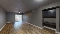 This single-story space is finished with a consistent, modern aesthetic throughout. Wood-look plank flooring runs across all living areas, complementing the cool gray wall tones and crisp white baseboards that define each room. The living area is generously proportioned, anchored by a ceiling fan and a sliding glass door fitted with vertical blinds, which allows natural light to filter in while providing access to an outdoor area with greenery views. A built-in shelving nook is integrated into one wall of the living space, offering functional display or storage potential. The open layout connects seamlessly toward the entry corridor, where a front door with a decorative transom window introduces additional natural light.

The kitchen is equipped with black appliances, including a range and over-the-range microwave, complemented by a mosaic tile backsplash and light countertops. A window above the kitchen area brings in further natural light. The bathroom features wood-look flooring, a tub-shower combo with tile surround, a vanity with a light countertop, and dark cabinetry. The bedrooms are well-sized, each with ceiling fans, and at least one includes a walk-in closet. A textured ceiling is present in select areas, adding subtle architectural character throughout the space.