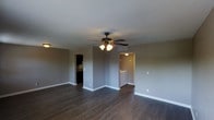 This single-floor residence offers a straightforward and functional layout designed for comfortable everyday living. The main living area is anchored by rich, dark hardwood-style flooring that runs throughout, providing a warm and cohesive aesthetic underfoot. Neutral grey walls create a clean, versatile backdrop well-suited for a variety of interior styles. A ceiling fan with an integrated light fixture is centrally mounted overhead, ensuring both air circulation and ambient lighting across the space. White baseboards provide a crisp contrast to the darker flooring, adding a polished finishing detail to the room. A dedicated opening leads to an in-unit laundry area, visible from the main living space and equipped with a washer and dryer, offering added convenience. An additional doorway provides access to a secondary room, likely a bedroom or utility space, partially visible with darker interior tones. The layout makes efficient use of the available footprint, with ceiling vents indicating a central HVAC system for year-round climate control. Overall, the space presents a clean, move-in-ready environment with practical features and a cohesive neutral color palette throughout.