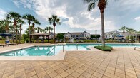 Located at 10558 Eastern Shore Blvd, this outdoor amenity area is centered around a generously sized, resort-style swimming pool with crystal-clear blue water. The pool deck is finished with elegant paver stonework that extends throughout the entire space, providing a clean and cohesive aesthetic. A separate wading or spa area is situated adjacent to the main pool, offering a tiered aquatic experience for residents.

The surrounding deck is well-furnished with multiple lounge chairs and seating arrangements, ideal for relaxation and sunbathing. Shade structures include at least one traditional wooden gazebo as well as a pergola-style covered seating area outfitted with patio umbrellas and comfortable outdoor furniture, providing ample options for shaded retreat.

Tall palm trees are strategically planted throughout the space, enhancing the tropical ambiance and providing natural shade. Manicured shrubs and green landscaping line the perimeter, adding a polished, resort-like feel. A black wrought iron fence borders the pool area, ensuring privacy and security while maintaining open sightlines to the surrounding multi-story residential buildings. The clubhouse building, clad in blue siding and brick accents, is directly accessible from the pool deck, seamlessly connecting indoor and outdoor amenities.