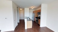 This single-floor residence offers a clean, contemporary layout designed for both functionality and comfort. The open-concept kitchen serves as the heart of the space, outfitted with a full suite of stainless steel appliances including a French door refrigerator, microwave, and range. Warm medium-toned wood cabinetry provides generous storage, complemented by dark stone countertops that add a touch of sophistication. A kitchen island with a built-in sink and lighter wood paneling creates a natural gathering point and additional prep space. Rich, dark hardwood-style flooring runs throughout, grounding the space with warmth and continuity. Track lighting above the kitchen adds focused illumination, while the open sightlines extend toward the entryway, which features a solid front door and a louvered closet door — ideal for additional storage. The white walls and neutral palette keep the interior feeling bright and airy. The layout flows intuitively, with clear visual connections between the kitchen and adjacent living areas, making the space practical for everyday living while maintaining a polished, modern aesthetic throughout.