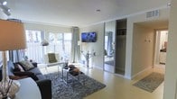 This single-floor space offers a thoughtfully designed open-concept layout that maximizes both functionality and style. The living area is anchored by light hardwood flooring and a plush area rug, creating a warm and inviting atmosphere. Large sliding glass doors flood the room with generous natural light while providing direct access to an outdoor patio, framed by lush greenery that enhances the sense of openness. Crown molding along the ceiling adds a refined architectural touch throughout the space. A wall-mounted flat-screen television serves as a focal point in the living area, complemented by a sleek glass console table and modern chrome accent furniture. The mirrored sliding closet doors are a standout feature, reflecting natural light and visually expanding the room, while also revealing a well-organized interior storage space. The neutral color palette of soft whites and warm beige tones creates a clean, contemporary ambiance that feels both airy and cohesive. A comfortable seating arrangement, complete with a dark sofa and a minimalist chrome coffee table set, ties the living space together elegantly. Overall, the space presents a modern, efficient, and well-lit environment ideal for comfortable everyday living.
