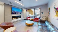 Located at 412 12th Ave SE in Minneapolis, MN, this single-floor communal amenity space is thoughtfully designed to offer a dynamic mix of relaxation, recreation, and productivity. The main lounge area serves as the heart of the space, anchored by a sleek linear gas fireplace set against tile surround, topped with a large wall-mounted flat-screen TV. Eclectic seating — including bold red leather accent chairs, a generous leather sofa, and colorful side tables — creates an inviting social atmosphere. Floor-to-ceiling glass windows flood the lounge with natural light and provide street-facing views of the Minneapolis neighborhood. Vibrant abstract artwork displayed on crisp white walls adds an energetic, creative tone throughout. Adjacent to the lounge is a game area centered around a full-size ping-pong table, complemented by a compact kitchenette featuring stainless steel appliances and mosaic tile backsplash, ideal for casual gatherings. A semi-private meeting nook, separated by a decorative laser-cut partition screen, offers a round table with soft green chairs — perfect for focused conversations or small group meetings. Track and pendant lighting throughout enhances the modern, polished aesthetic of the entire space.