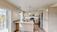 Located at 13808 E Carlisle Ave in Spokane Valley, WA, this single-floor unit offers a clean, contemporary design with a neutral color palette throughout. The open-concept kitchen serves as a central highlight, showcasing light maple cabinetry with brushed nickel hardware, quartz countertops, and a full suite of stainless steel appliances including a range, over-the-range microwave, and refrigerator. A spacious central island with an undermount sink provides additional prep space and casual seating, while two pendant lights with metallic finishes hang above, complementing the recessed lighting overhead. Light wood-tone luxury vinyl plank flooring extends through the main living areas, adding warmth and continuity. The living area benefits from oversized windows, including a stacked window arrangement that invites abundant natural light and frames views of surrounding trees. Bedrooms feature patterned carpet flooring and large windows overlooking the Spokane Valley landscape. The bathroom is well-appointed with light wood cabinetry, a wide quartz vanity, a large mirror, and a tall linen tower for storage. An in-unit closet with built-in shelving and a dedicated entryway with louvered closet doors complete this thoughtfully designed space.