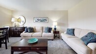 This single-floor residence offers a thoughtfully designed layout that flows naturally between living, dining, kitchen, and sleeping spaces. The kitchen is outfitted with white shaker-style cabinetry, granite-look countertops, a subway tile backsplash, and a full suite of stainless steel appliances, including a refrigerator, dishwasher, and electric range. Wood-look flooring grounds the kitchen with a warm, durable finish. Adjacent to the kitchen, the dining area features neutral carpet, a wall-mounted air conditioning unit, a baseboard heater, and a double window that brings in natural light and outdoor views. The living room is a comfortable, carpeted space with neutral walls and understated décor, creating a calm and inviting atmosphere. The bedroom is generously sized, with plush carpet, a large window overlooking greenery, and access to a closet. It connects directly to a bathroom finished with white tile wainscoting, wood-look flooring, a vanity with white cabinetry, and clean, minimal fixtures. Throughout the residence, a consistent neutral color palette and practical finishes contribute to a cohesive, functional living environment.
