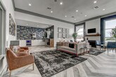 This thoughtfully designed single-story space functions as a polished community or leasing hub, blending contemporary aesthetics with functional amenities. The entry is anchored by a prominent reception desk with a warm wood-topped counter, white paneled base, and pendant lighting, all set against deep charcoal accent walls. Herringbone-patterned wood-look flooring runs continuously throughout, lending cohesion and visual texture to the entire space.

The open lounge area offers generous seating arrangements, including a tufted sofa, leather armchairs, and bistro-style tables with navy blue chairs, all positioned atop richly patterned area rugs beneath elevated ceilings with recessed lighting and exposed wood beam accents. A dedicated sitting area features a tiled fireplace with a dark wood mantel, a wall-mounted television, large-scale geometric artwork, and a picture window that invites natural light and outdoor views.

Private rooms with glass-paneled doors provide quieter settings for meetings or individual use. A beverage station serves as a standout feature, showcasing an eye-catching patchwork blue tile backsplash, white cabinetry with brass hardware, a built-in beverage cooler, and a specialty coffee machine. The overall ambiance is modern, curated, and welcoming.