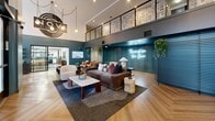 Located at 321 2nd Ave W in Seattle's Queen Anne neighborhood, Novi at Queen Anne presents a thoughtfully designed single-floor leasing office that blends contemporary aesthetics with functional workspace. The entry lobby commands immediate attention with soaring double-height ceilings, floor-to-ceiling steel-framed glass windows that flood the space with natural light, and striking herringbone-patterned hardwood flooring throughout. Deep teal accent walls serve as a bold design anchor, complemented by vibrant music-themed artwork, a glowing neon sign reading "This Must Be the Place," and a mezzanine display of framed cassette tapes that reinforces a distinct cultural character. The main lounge area is furnished with plush leather sofas, rounded accent chairs, and marble-topped coffee tables, creating an inviting and comfortable atmosphere. Globe pendant lights and brass wall sconces add warmth to the open layout. A dedicated conference room features an elegant oval brass-accented table, designer chairs, and a wall-mounted display, ideal for private meetings. Two private offices are finished with carpeted flooring, teal accent walls, and ample desk space. Lush indoor plants are distributed throughout, enhancing the fresh, welcoming ambiance of this well-appointed professional space.