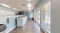 This single-floor laundry facility is designed to accommodate a high volume of users efficiently and comfortably. The space is finished with durable wood-look flooring and painted in a calming light blue throughout, creating a clean and welcoming atmosphere. Fluorescent lighting runs across the textured white ceiling, ensuring the entire room is evenly and brightly illuminated.

Two rows of full-sized top-load washing machines are arranged along the walls, complemented by a matching bank of stacked dryers, providing ample capacity for simultaneous use. A continuous white folding counter runs alongside the machines, offering a convenient surface for sorting and folding laundry. A Coca-Cola vending machine is positioned along one wall for added convenience.

Large French-style glass doors and multiple windows along one side of the facility allow natural light to filter in and provide views of the surrounding exterior. A seating area with white chairs is positioned near the entrance, giving users a comfortable place to wait. Trash receptacles are thoughtfully placed throughout the space. The overall layout is open and spacious, allowing for easy movement and a practical, functional experience.