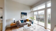 This single-floor residence showcases a contemporary design aesthetic characterized by elevated ceilings and a neutral, light-filled palette throughout. The living area is a standout feature, anchored by dramatic floor-to-ceiling windows and a full-height sliding glass door that opens directly onto a private balcony, offering urban views and an abundance of natural light. The generous ceiling height amplifies the sense of openness in the space.

The kitchen is thoughtfully designed with a clean, modern sensibility. A combination of warm wood-toned lower cabinetry and high-gloss white upper cabinets extends to the ceiling, maximizing storage. Light blue glass tile forms a refined backsplash, complemented by white quartz countertops, a kitchen island, and a full suite of stainless steel appliances including a range, dishwasher, built-in microwave, and refrigerator. Light hardwood flooring runs throughout the main living areas, enhancing warmth and continuity.

The bedroom benefits from tall windows that frame outdoor views while allowing natural light to fill the carpeted room. The bathroom features wood-grain vanity cabinetry with a white quartz countertop, an illuminated mirror, and a tub-shower combination with tile flooring, completing the space with a polished, functional finish.