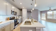 This single-floor residence showcases a thoughtfully designed open-concept layout with a cohesive, contemporary aesthetic throughout. The kitchen serves as a central highlight, featuring gray shaker-style cabinetry, white quartz countertops, and a full suite of stainless steel appliances including a gas range, over-the-range microwave, and refrigerator. A generously sized center island with an undermount sink, seating for two, and pendant lighting above creates an ideal space for both meal preparation and casual dining. Recessed lighting and clear glass pendant fixtures add layers of illumination across the space.

Light wood-style flooring flows continuously from the kitchen into the open living and dining areas, which benefit from natural light through large windows and a glass door. A ceiling fan enhances comfort in the living area. The bedrooms are well-proportioned, featuring neutral tones, ceiling fans, and windows that invite natural light. The bathroom is finished with a white quartz vanity top, undermount sink, large wall mirror, and a tub-shower combination with tile surround. Soft, muted wall tones in lavender-gray and warm taupe unify the space, creating a calm and modern residential atmosphere throughout.