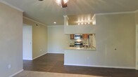 This single-floor residence is designed with an open-concept layout that seamlessly connects the living area and kitchen. The living space features rich, dark hardwood-style flooring, white baseboards, and crown molding that frame the room with clean, classic detail. A ceiling fan with a light fixture is centrally positioned overhead, providing both air circulation and ambient lighting.

The kitchen is defined by a raised pass-through counter with granite countertops, creating a natural divide between the cooking and living spaces while maintaining an open, connected feel. The kitchen is equipped with stainless steel appliances, including a range, microwave, and refrigerator, complemented by warm wood-toned cabinetry. Track lighting mounted above the kitchen illuminates the workspace effectively.

Decorative columns with crown molding detail frame the kitchen pass-through, adding an architectural focal point to the space. The neutral, warm wall tones throughout contribute to a cohesive and inviting atmosphere. A doorway visible on the left side of the living area suggests additional rooms beyond, completing this well-appointed single-level residence.