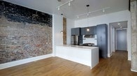 This single-floor space masterfully combines industrial character with contemporary finishes. The open layout is anchored by striking exposed brick walls, which lend a raw, textural quality that contrasts beautifully with the clean, modern aesthetic throughout. Warm-toned hardwood flooring runs continuously underfoot, adding natural warmth to the space.

The kitchen is a standout feature, designed with dark gray flat-front cabinetry that extends to the ceiling for ample storage. Stainless steel appliances, including a refrigerator and gas range, are complemented by a white geometric tile backsplash. A large white waterfall island serves as the central workspace and gathering point, complete with an integrated sink.

Lighting is thoughtfully layered, combining adjustable track lighting mounted on the ceiling with two globe pendant lights suspended above the island, creating both functional and ambient illumination. The high ceilings enhance the sense of openness and volume, contributing to an airy, loft-like atmosphere. Overall, the space offers a refined yet edgy aesthetic, ideal for those who appreciate the blend of historic character and modern convenience.