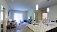 This single-floor residence offers a thoughtfully designed open-concept layout that seamlessly connects the kitchen, dining, and living areas into one cohesive and functional living space. The kitchen anchors the layout with a striking granite-topped island that doubles as a breakfast bar and entertaining surface, illuminated by a sleek pendant light overhead. The dining area flows naturally from the kitchen, offering a comfortable setting for meals and gatherings. The living room features plush carpeting and a large window dressed with vertical blinds that allows natural light to fill the space, creating a bright and welcoming atmosphere. A television console with decorative cabinetry serves as the focal point of the living area. The neutral warm-toned walls throughout contribute to a calm and cohesive aesthetic. A hallway visible from the main living area leads to the private bedroom, which features a glass-paneled door that adds a touch of elegance and allows borrowed light to travel through the space. The stainless steel refrigerator is partially visible, hinting at a fully equipped kitchen. Overall, the space presents a clean, modern, and comfortable environment well-suited for everyday living.