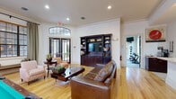 This single-floor community amenity space is thoughtfully designed to balance relaxation, entertainment, and fitness. The main lounge area serves as the centerpiece, featuring warm hardwood flooring, crown molding, and generous natural light streaming through large windows dressed with elegant drapes and blinds. Comfortable leather seating surrounds a stylish coffee table, while a built-in dark wood entertainment unit with a flat-screen TV anchors one wall. A full-size billiards table with pendant lighting overhead adds a social focal point to the room. Seating nooks near the windowed walls and French doors leading to the exterior create an open, inviting atmosphere. The kitchen is well-equipped with dark espresso cabinetry, stainless steel appliances including a refrigerator and microwave, a vibrant mosaic tile backsplash in warm red tones, and a long breakfast bar counter. Adjacent to the lounge, the fitness center is divided into distinct workout zones. One area houses cycling machines, a power tower, and a rowing mat, while a second zone features a full kettlebell rack, medicine ball storage, and wall-mounted TV — all surrounded by large windows overlooking lush greenery. The consistent use of light walls, natural wood tones, and quality finishes throughout creates a cohesive and welcoming environment.