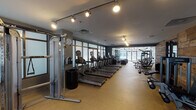 This fitness center is a comprehensively equipped facility designed to accommodate a wide range of workout styles and fitness goals. The space is organized into distinct functional zones, each purpose-built for specific types of training.

The cardio area features a substantial row of treadmills and elliptical machines, each equipped with individual digital screens. Large floor-to-ceiling windows line the perimeter, flooding the space with natural light and offering outdoor views, creating an open and energizing environment. Track lighting mounted on the ceiling provides additional illumination throughout.

The strength training section is well-stocked with multiple cable and weight machines, a rack of dumbbells, barbells with weight plates, and adjustable benches—offering thorough options for resistance training. Rubber flooring throughout this zone ensures both safety and durability.

A dedicated studio area features smooth hardwood-style flooring, a wall-mounted flat-screen television framed in reclaimed wood, and exercise balls, making it well-suited for stretching, yoga, or guided workouts. A striking reclaimed wood accent wall adds a warm, rustic design element that contrasts effectively with the facility's otherwise contemporary, gray-toned aesthetic.

A separate alcove houses additional treadmills alongside frosted glass interior partitions, offering a quieter, semi-private cardio option. Overall, the facility presents a polished, modern environment with thoughtful design and a comprehensive equipment selection.