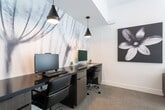 Located at 26451 Camino de Vista in San Juan Capistrano, CA, this single-floor commercial space combines modern design with functional workspaces in a cohesive, contemporary aesthetic. The interior is defined by vaulted ceilings that lend an open, airy feel throughout, complemented by neutral tones, plush carpeting, and an abundance of natural light filtering through large roller-shaded windows.

The business center is a standout feature, anchored by a long dark wood desk accommodating two workstations, each equipped with a computer and ergonomic chair. A dramatic large-scale botanical mural serves as a striking backdrop, while a row of sleek pendant lights overhead adds focused illumination. A bold black-and-white floral canvas artwork further enhances the artistic character of this area.

Adjacent to the business center, a lounge area provides two plush dark armchairs paired with a small side table, positioned near a windowed exit door that offers views of surrounding greenery — ideal for informal meetings or relaxed work sessions.

A dedicated booth seating area features upholstered banquette-style benches with white tabletops and pendant lighting above, creating semi-private spaces well-suited for collaboration or casual gatherings. Framed artwork adorns the walls throughout, reinforcing the space's refined, design-forward atmosphere.