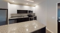 This well-appointed single-floor residence offers a clean, contemporary design throughout. Dark hardwood-style flooring flows seamlessly across the open-concept living and dining area, creating a cohesive and polished aesthetic. Neutral-toned walls complement the light-filled layout, with a window in the living space allowing natural light to brighten the interior.

The kitchen is a standout feature, showcasing sleek white upper cabinetry contrasted by dark lower cabinets, granite countertops with subtle speckling, and a matching granite-topped island ideal for dining or meal prep. Stainless steel appliances — including a refrigerator with ice and water dispenser, electric range, built-in microwave, and dishwasher — complete the modern culinary space. Recessed lighting overhead adds a warm, functional glow.

A hallway connects to multiple rooms, including bedrooms and utility spaces, all finished with consistent flooring and neutral paint. The bathroom features a full tub-shower combination with chrome fixtures and tile flooring. Conveniently, a stacked washer and dryer unit is located within the unit, accompanied by wire shelving for added storage. The layout is practical and efficient, making excellent use of its footprint for comfortable everyday living.