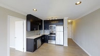 This single-floor space showcases a well-appointed kitchen that serves as a focal point of the layout. The kitchen features a striking contrast between dark espresso cabinetry and light countertops, creating a bold, contemporary aesthetic. Upper and lower cabinets provide generous storage, while a grey subway tile backsplash adds texture and visual interest to the cooking area. The kitchen is equipped with stainless steel appliances, including a built-in microwave, electric range, and dishwasher, alongside a white refrigerator. Recessed and track lighting illuminate the workspace efficiently from above. Light-toned hardwood flooring extends throughout the space, enhancing the bright and open feel of the interior. A hallway visible beyond the kitchen suggests additional rooms, connecting the space cohesively. Crown molding and clean white trim provide refined architectural detailing, while the open layout between the kitchen and adjoining areas promotes a seamless flow. The overall design balances functionality with modern style, making it a practical and visually appealing living environment.