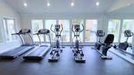 This fitness center offers a clean, modern workout environment designed to support an active lifestyle. The space is anchored by a row of large windows that span the front wall, flooding the room with natural light and providing open views of the outdoor greenery and surrounding area beyond. The bright, airy atmosphere is further enhanced by recessed ceiling lighting, ensuring consistent illumination throughout.

The gym is outfitted with a thoughtfully arranged selection of cardio equipment, including treadmills, elliptical cross-trainers, and a stationary bike. The machines are positioned to face the expansive windows, allowing users to enjoy the outdoor scenery during their workouts. The flooring is finished with a durable, dark rubberized surface that is both practical and easy to maintain, providing a safe and stable foundation for exercise.

The walls are finished in a crisp white, contributing to the space's clean and contemporary aesthetic. The overall layout is open and uncluttered, maximizing usable floor space and allowing for comfortable movement between equipment. The combination of natural light, outdoor views, and well-maintained equipment makes this fitness center a functional and inviting space for daily exercise.