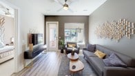 This single-story space showcases a cohesive modern design with a neutral color palette of warm taupes and cool grays carried throughout. The living area serves as the heart of the unit, featuring light wood-style flooring, a ceiling fan, and large windows alongside a glass-panel door that invite natural light and provide views of the outdoor surroundings. A bold accent wall anchors the space, complemented by sculptural wall art and mid-century modern furnishings that lend a curated, contemporary feel.

The kitchen is well-appointed with flat-front gray cabinetry, stainless steel appliances including a range, microwave, refrigerator, and dishwasher, as well as a white subway tile backsplash and light quartz countertops. A spacious island with pendant lighting above offers additional prep space and casual seating.

The bedroom continues the warm neutral tones with plush carpeting, a ceiling fan, a window with outdoor views, and distinctive branch-inspired wall décor that adds organic texture. Ample closet access is provided via multiple doors off a short hallway.

The bathroom features large-format tile flooring, a single-sink vanity with dark wood cabinetry, and a tub-shower combination, maintaining the clean, modern aesthetic found throughout the space.