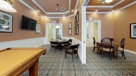 This sophisticated recreational lounge is designed to offer a refined yet relaxed environment for gaming and socializing. The space flows across a single floor, with distinct areas seamlessly connected through wide doorways and French doors that lend an open, airy feel throughout.

The game room areas are anchored by a full-size billiards table set beneath a wrought-iron pendant light fixture, complemented by a wall-mounted flat-screen television and a dedicated cue rack. Natural light filters in through large arched windows dressed with ornate valances, enhancing the warm, inviting atmosphere.

Multiple card and game tables — including an octagonal table ideal for poker or board games — are accompanied by elegantly crafted wooden chairs with lattice-back detailing and upholstered seating. The rooms are finished with rich, warm-toned walls, white wainscoting, and tray ceilings with crown molding that add a classic architectural charm.

Throughout, a bold geometric-patterned carpet in navy and gold ties the spaces together with a touch of elegance. Framed artwork and decorative accents complete the polished aesthetic. French doors open to additional areas, suggesting seamless access to further amenities, making this lounge a well-rounded destination for leisure and entertainment.