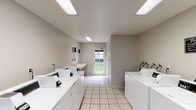 This single-floor shared laundry facility is designed for practicality and efficiency, offering a spacious, well-organized layout suited to accommodate multiple users simultaneously. The room features rows of commercial-grade top-loading washing machines and dryers lining both sides of the space, providing substantial capacity for high-volume use. Each machine is equipped with individual payment terminals, streamlining the laundry process for users.

The flooring throughout consists of light-toned ceramic tiles, contributing to a clean and easy-to-maintain environment. Neutral gray walls keep the aesthetic simple and functional, while flush-mounted fluorescent light panels provide consistent, bright illumination across the entire room. A small window along one wall and a glazed exterior door at the far end allow natural light to filter in, offering views of the surrounding outdoor greenery.

A built-in countertop workspace is positioned near the entrance, providing a convenient surface for folding or sorting laundry. Instructional signage is posted on the walls, offering guidance on machine operation and laundry room rules. The room's straightforward, uncluttered design prioritizes ease of use, making it a highly functional communal amenity within its broader property setting.